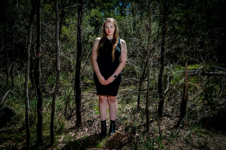 A woman with long brown hair, in black dress stands in partially burnt out bush, stark black and green colours. 