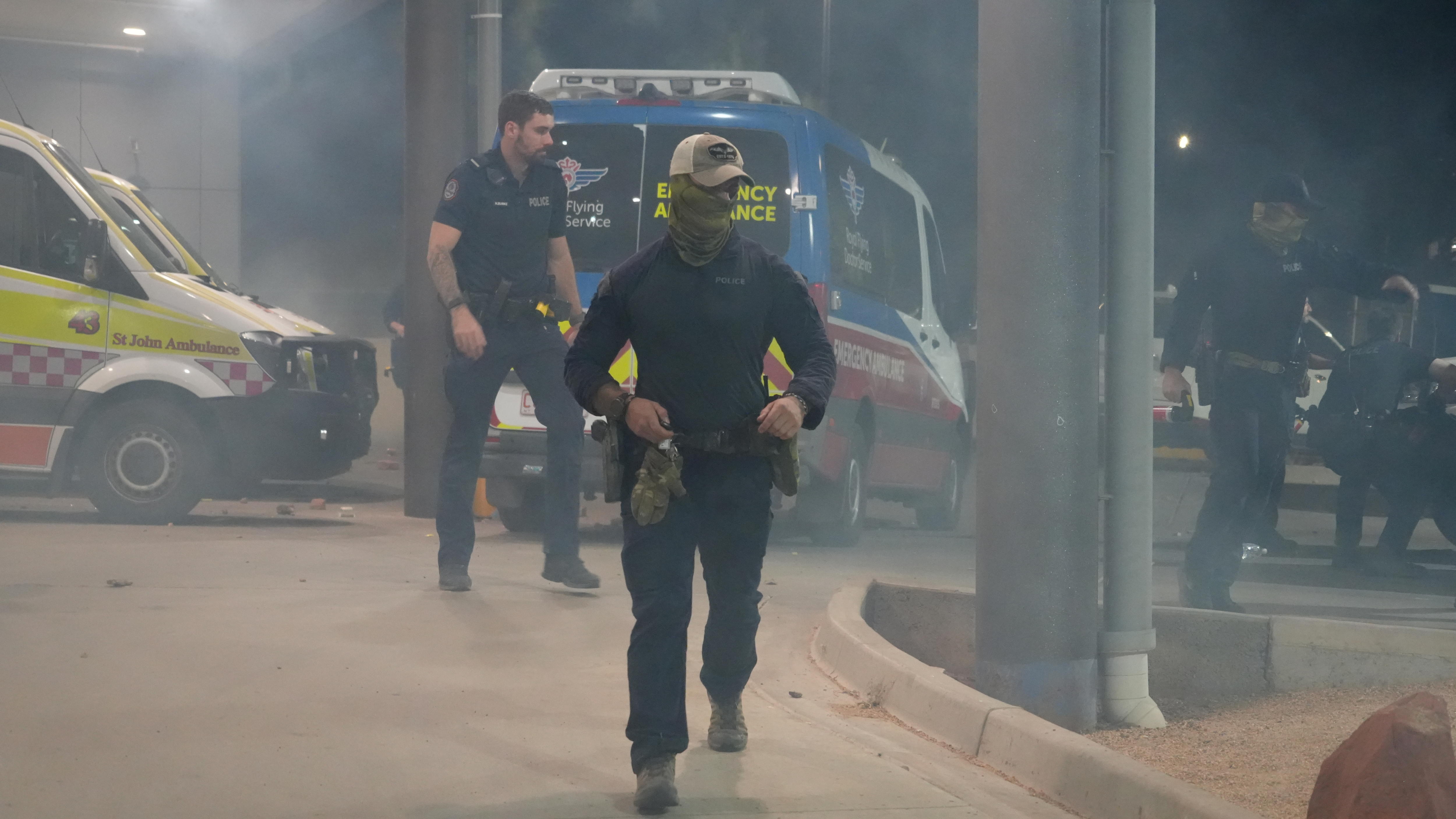 Un oficial de policía lleva un pañuelo sobre la boca y la nariz y una gorra de béisbol en la rampa de una ambulancia en el hospital.