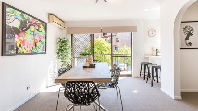 A large dining table is seen near the balcony of a brightly lit inner suburb apartment