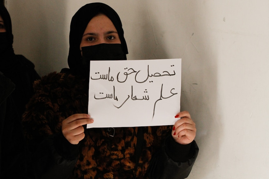 A woman wears a hijab and face covering while holding a protest sign with two hands.