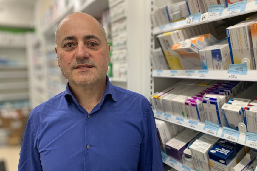 Angelo wearing a blue business shirt, standing in front of shelves full of prescription medications.
