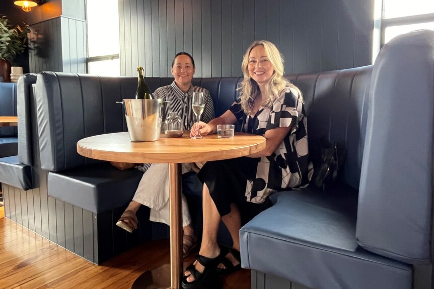 Two women in a blue booth at a restaurant with a champagne bottle and glasses