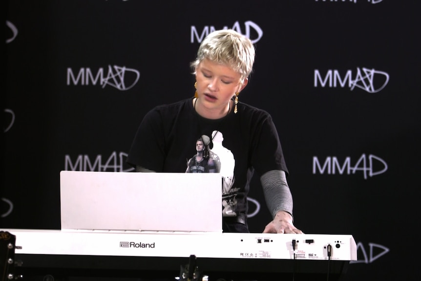 A woman with short blonde hair plays a keyboard with a black backdrop printed with MMAD 