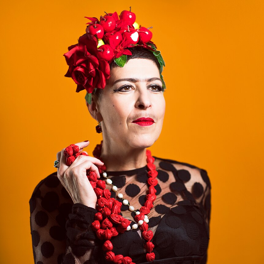 Natalie Gamsu as Carmen stands in front of an orange backdrop wearing a black dress and a hat with fruit and a rose attached.