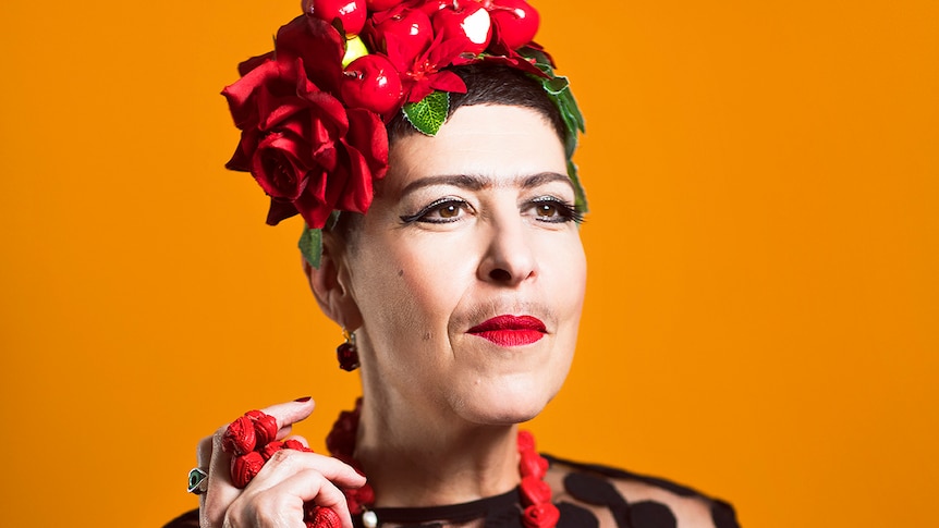 Natalie Gamsu as Carmen stands in front of an orange backdrop wearing a black dress and a hat with fruit and a rose attached.