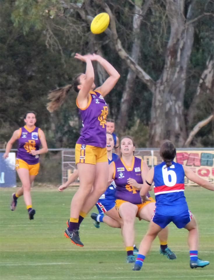 A woman in a purple and yellow football uniform leaps up to mark the ball