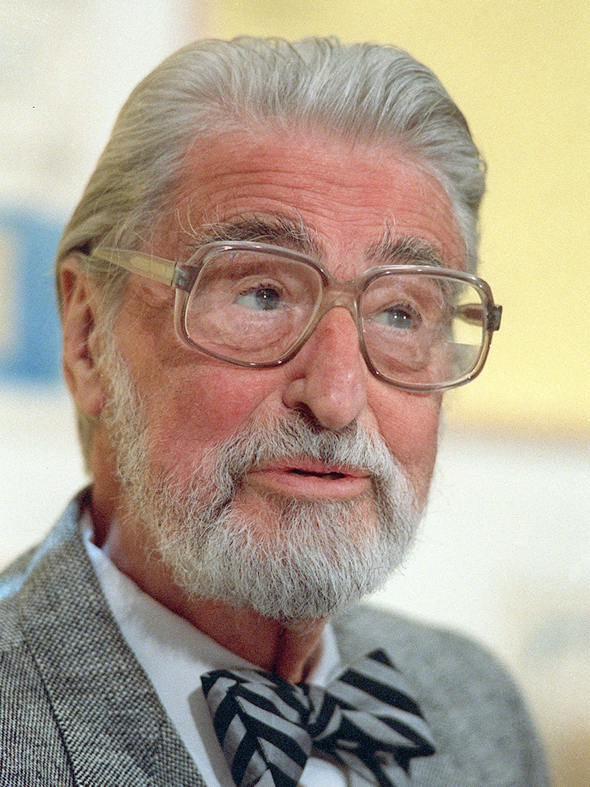 A man in a grey suit and glasses pauses as he speaks.