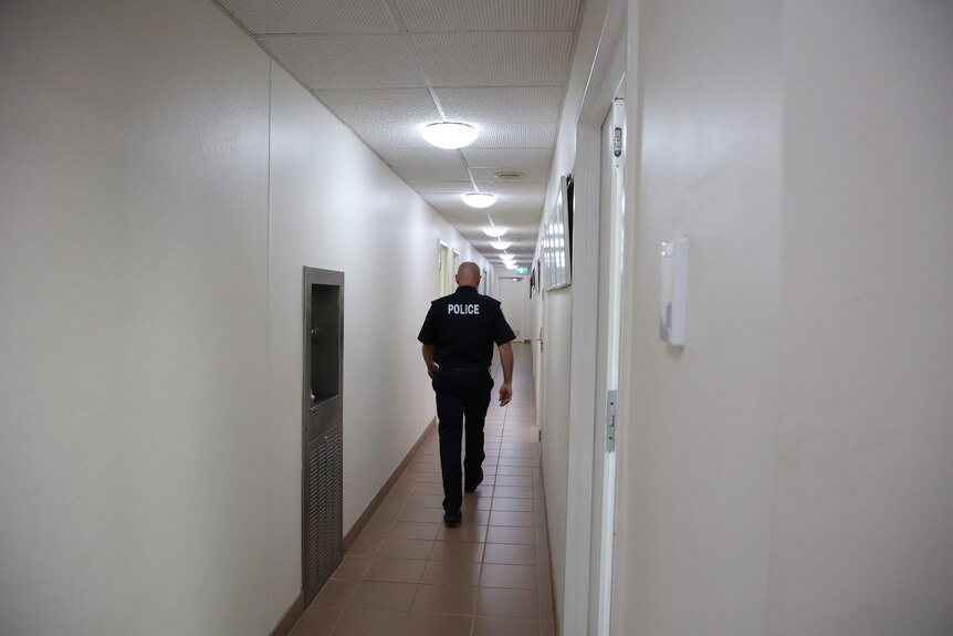 A Northern Territory police officer walks down a hallway with bright white lighting.