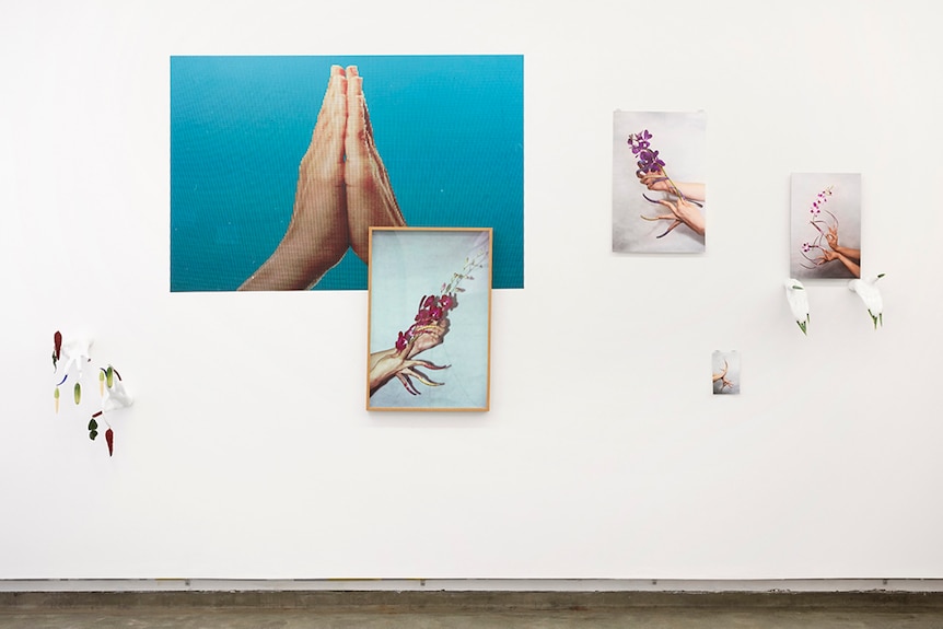 Photos of hand gestures and white plaster sculptures of hands with fake nails and plastic fruit displayed on white gallery wall.