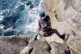 Gideon Mettam perches on a ledge on a sea cliff with a rope for safety.