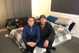 An image of Kelly and her partner Daniel sitting on a mattress on a floor with packed bags behind them