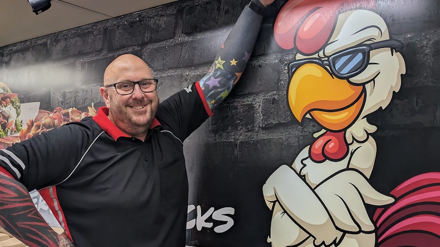 bald man with glasses with left arm raised against a wall with cartoon chicken painted on it
