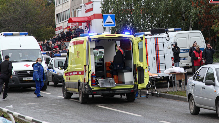 A yellow ambulance with its back doors open as more emergency service vehicles and workers move around it. 