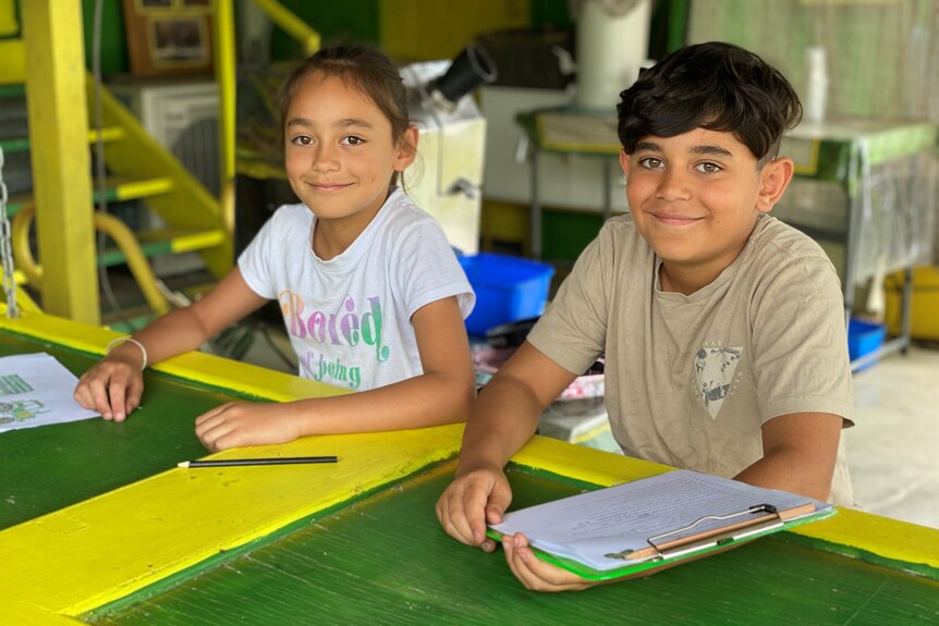 A girl and boy sitting at a table in a yellow and green table with a notebook in hand.