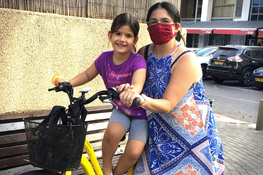 A woman in a face mask sits on a bike with a small girl
