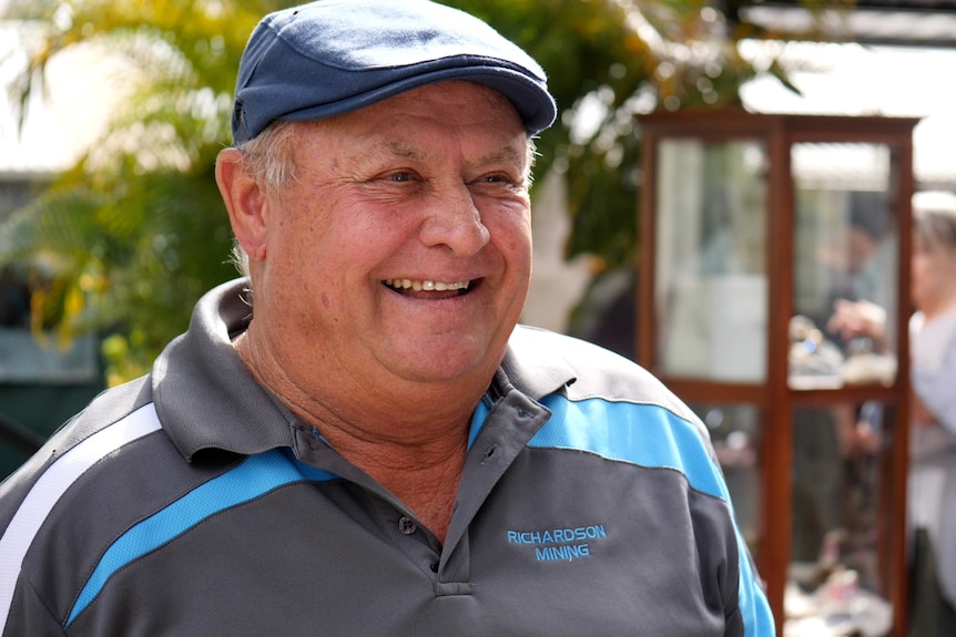 Ray wearing a small hat smiling, wearing polo shirt, jewellery cabinet behind.