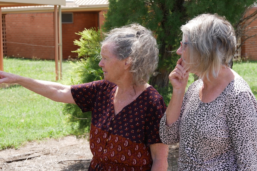 Two elderly women standing outdoors look away from camera and point to something in the distance. 