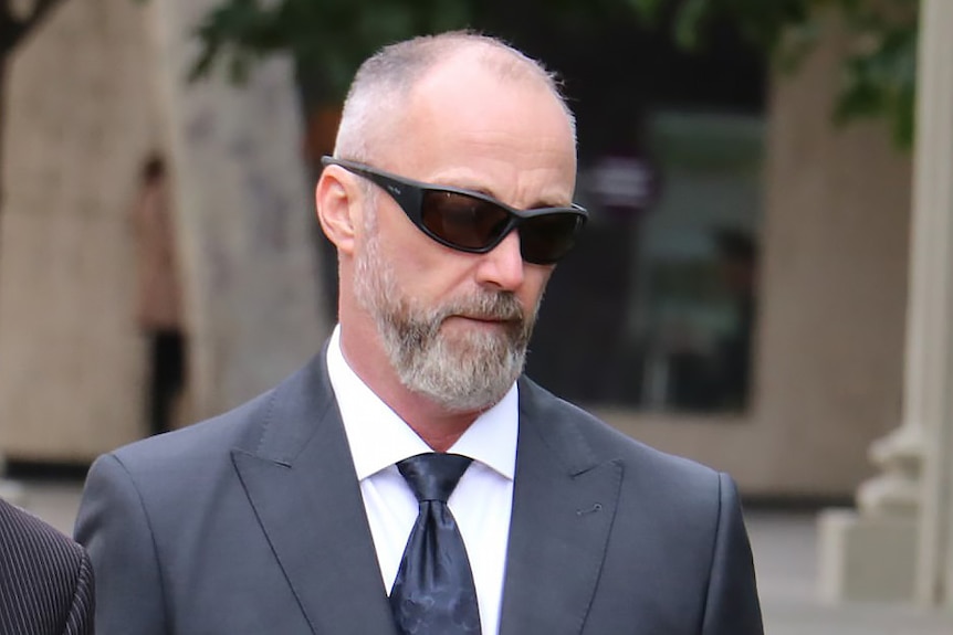A close-up shot of a balding man with a grey beard and black sunglasses looking downwards outside, wearing a suit and tie.