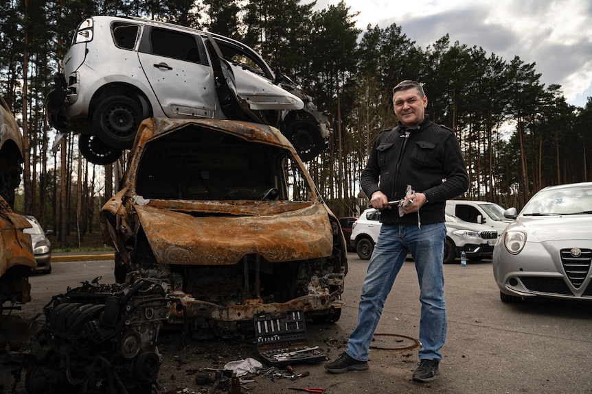 A man holding tools stands next to two cars: a damaged silver hatchback sits precariously atop the burned hull of another car