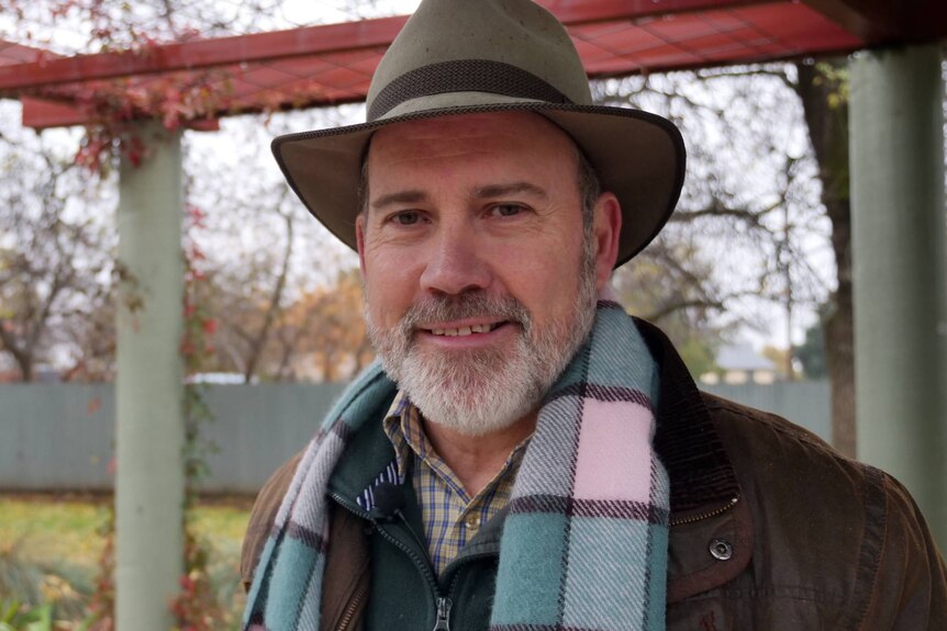 A man in a hat and scarf smiles at the camera with a park in the background.
