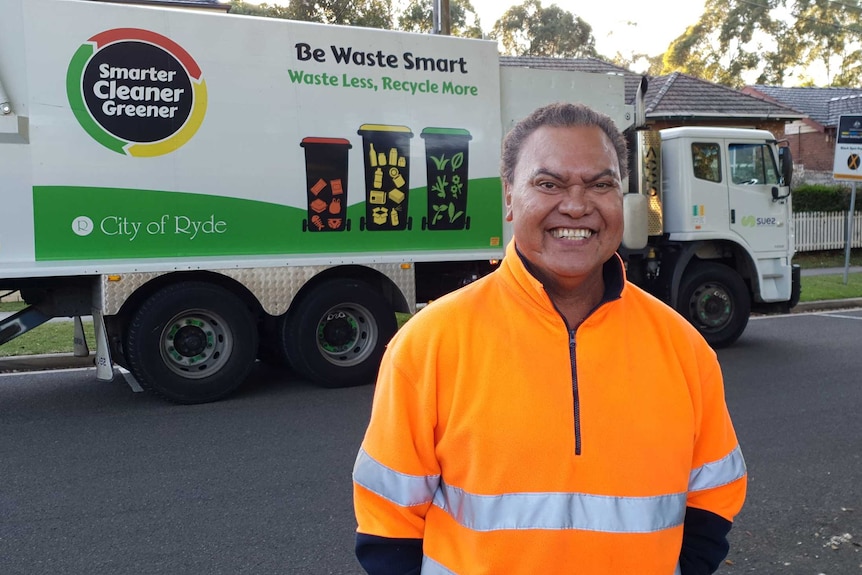 A smiling Olsen Filipaina stands in front of his rubbish truck.
