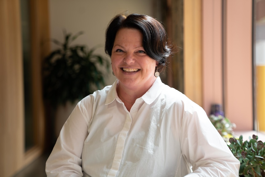 Natalie Foley, a woman with short dark brown hair and who's wearing a white shirt, poses for a photo, smiling, at a sunlit bench