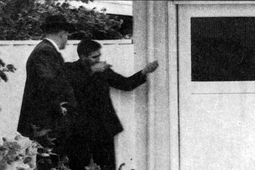 Eric Edgar Cooke mimes shooting a rifle outside a house as a detective watches.
