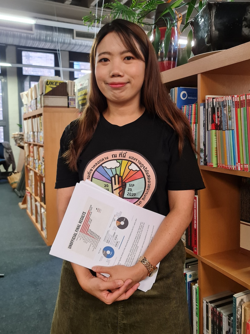 You view a Thai woman with brown hair standing in front of a bookshelf in an open office.