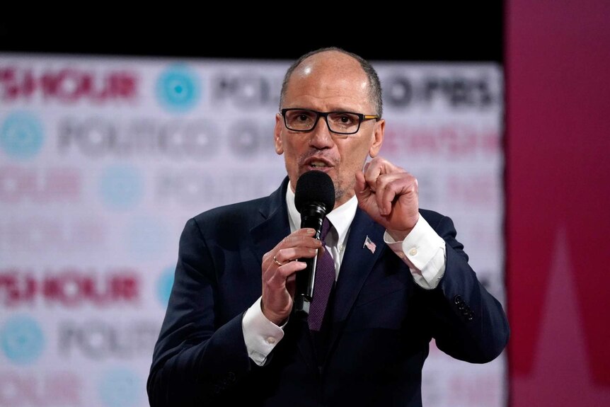 Tom Perez in glasses and a suit holding a microphone and holding his hand up in front of him with an out of focus sign behind