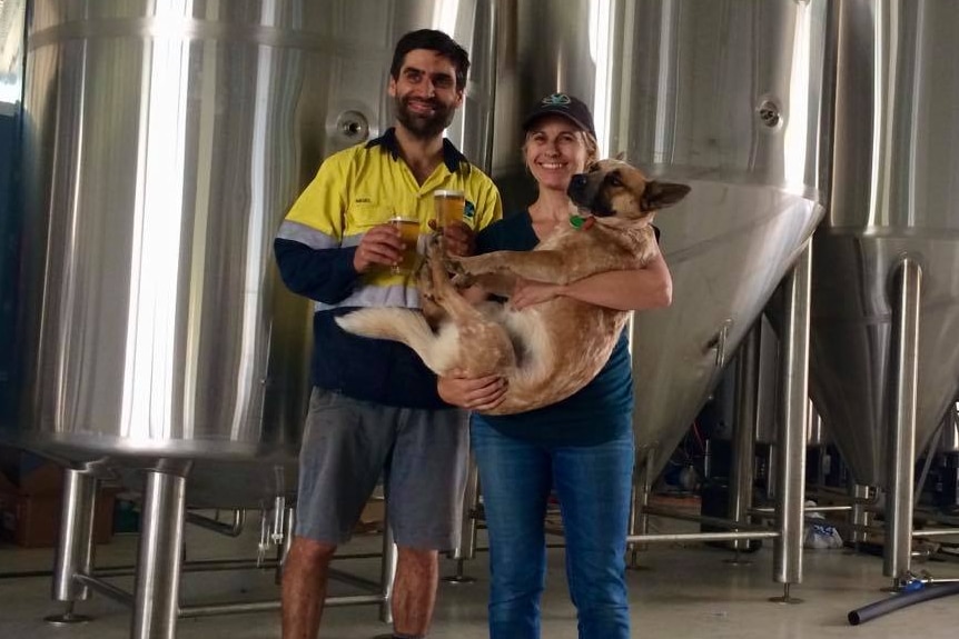 A man and a woman, both with big smiles, stand in front of brewing equipment. The woman is cradling a large dog.