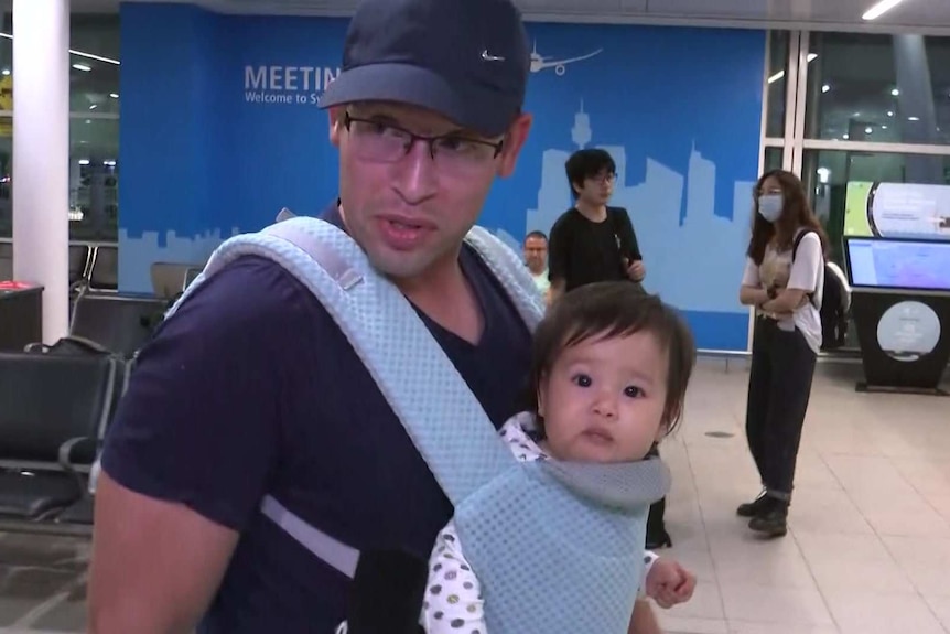 A man carries his young daughter through the airport in a baby carrier on his chest, with passengers wearing masks behind them.