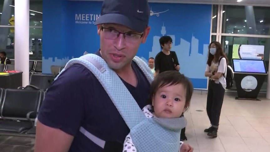 A man carries his young daughter through the airport in a baby carrier on his chest, with passengers wearing masks behind them.