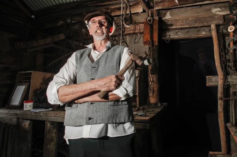 A man with grey hair wear a cap and a vest, holding a hammer in an maritime village shop