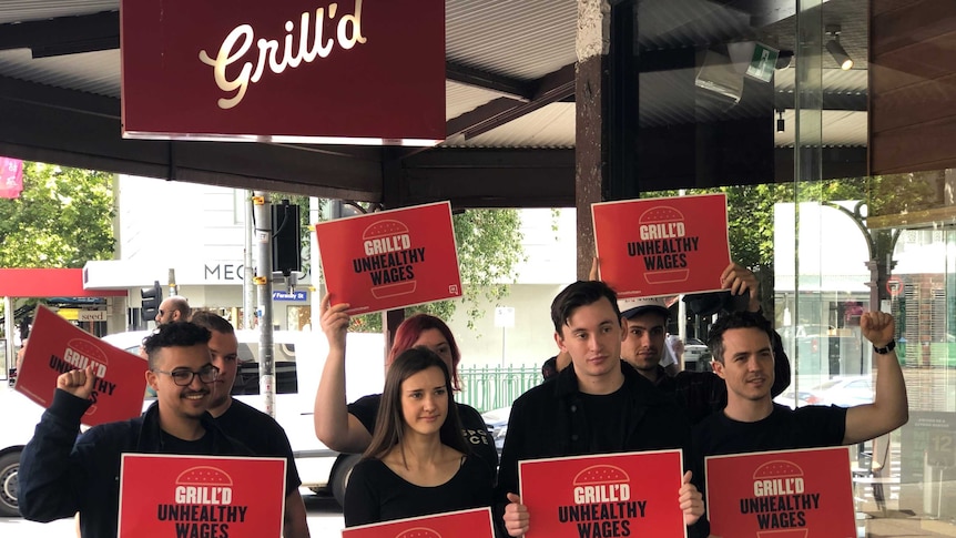 A group of young people wearing black hold red signs saying 'Grill'd unhealthy wages'.