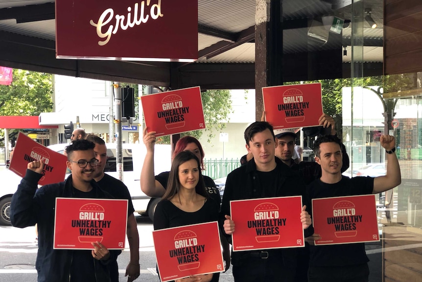 A group of young people wearing black hold red signs saying 'Grill'd unhealthy wages'.