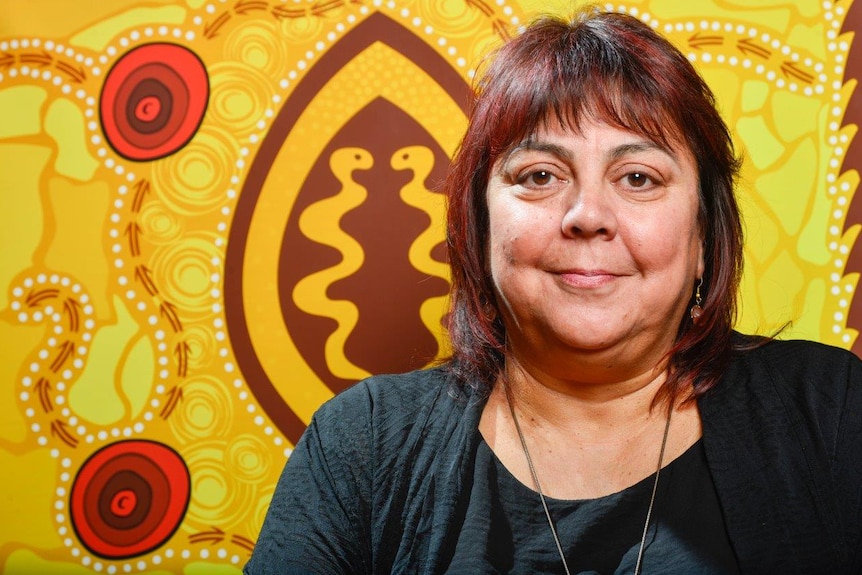 A woman stands in front of a yellow Indigenous artwork.