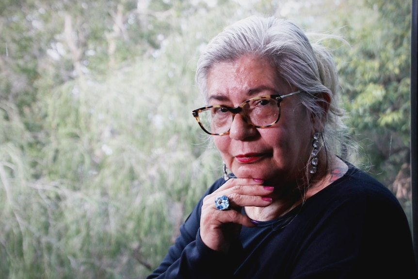 A woman with white hair and glasses touches her chin with her fingers.