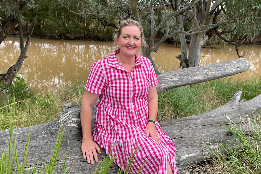 A lady with blonde hair in a pink and white checkered dress sitting on a log smiling with a river in the background