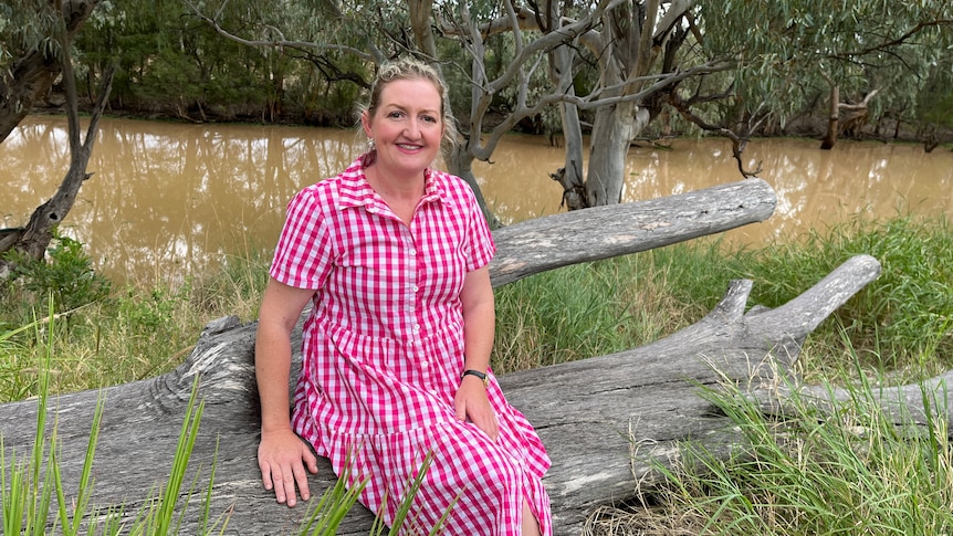 A smiling blonde woman in a checked dress sits on a fallen tree in front of a river.