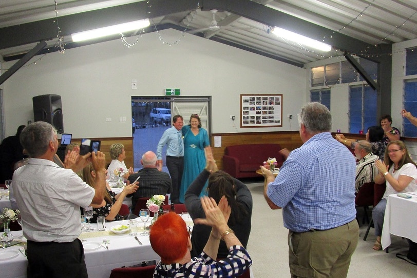 Newly married couple walks into large shed with people sitting at tables clapping and cheering.