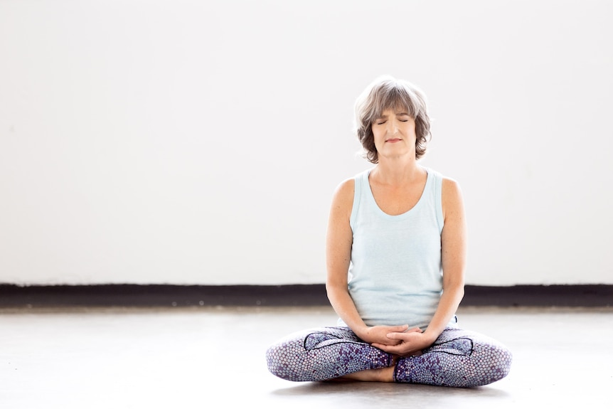 An older fit woman with short grey hair sits cross legged on the ground meditating in a room.
