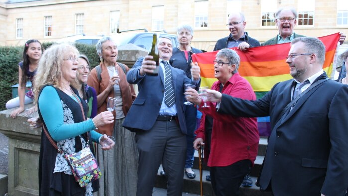 Rodney Croome surrounded by people pops champagne.