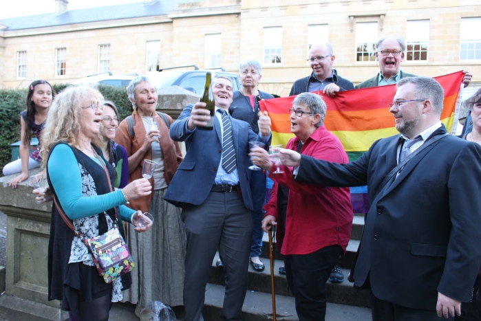 Rodney Croome surrounded by people pops champagne.