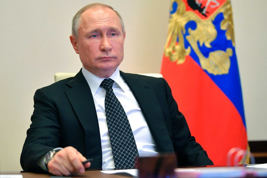 President Vladimir Putin stares as he sits in front of a lag behind a desk.