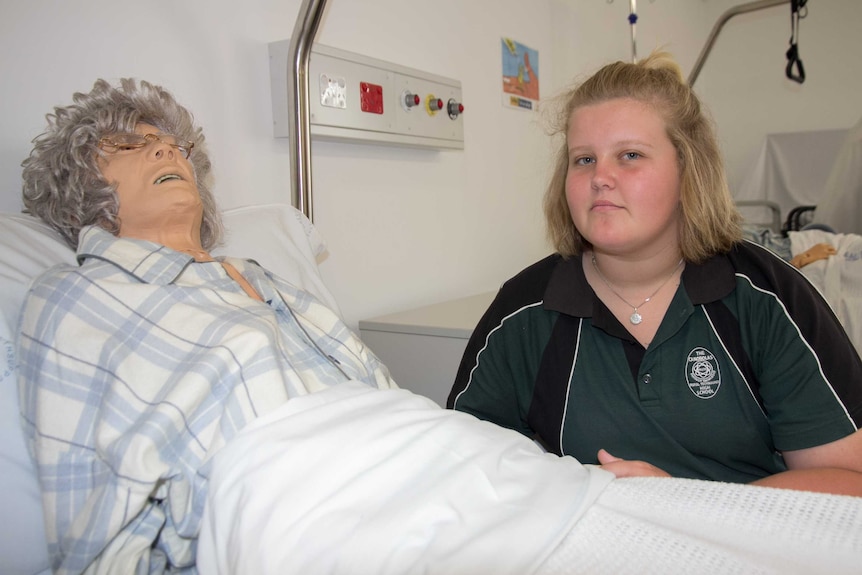 A teenager in a school shirt sitting near a hospital bed with an elderly mannequin