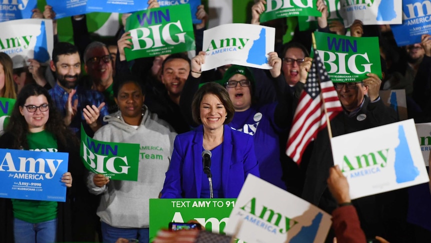 Amy Klobuchar smiling at a podium surrounded by supporters with "win big" signs