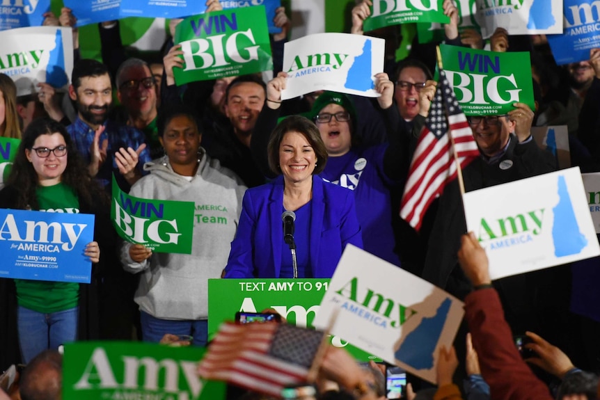 Amy Klobuchar smiling at a podium surrounded by supporters with "win big" signs