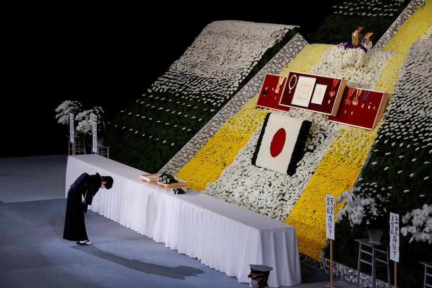 A woman in dark formal clothes bow deeply towards a large memorial shrine with yellow and white flowers and the Japanese flag.