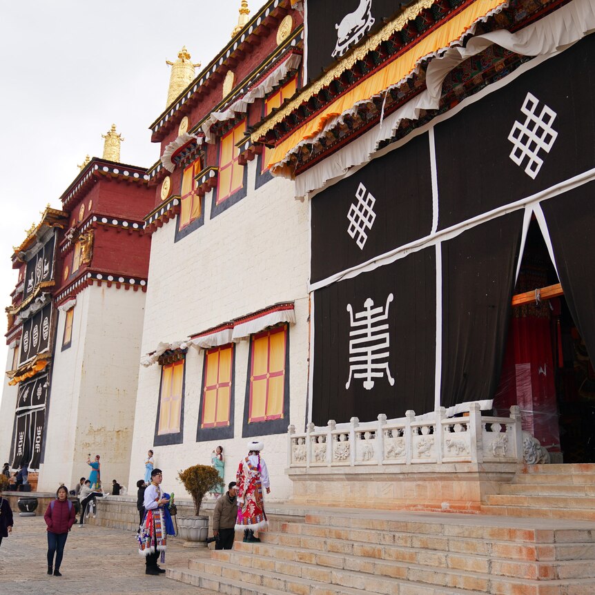  Tourists at the Ganden Sumtsenling Monastery, a large light coloured building with large black cloth with symbols at entrance