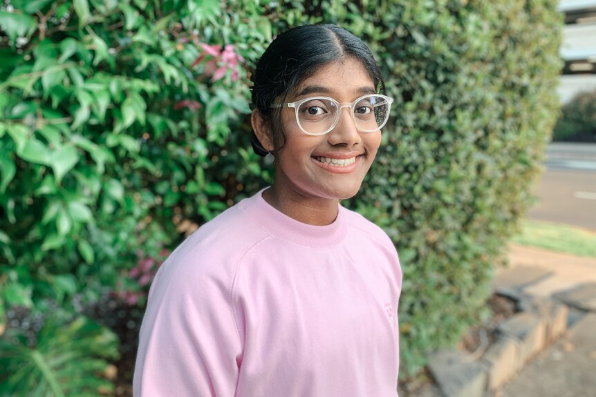 A young girl with brown skin, a pink sweater and glasses smiles for the camera in front of a backdrop of plants.